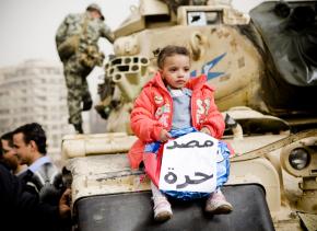 A child holds a sign saying "Egypt is Free" while sitting on a tank parked in Tahrir Square